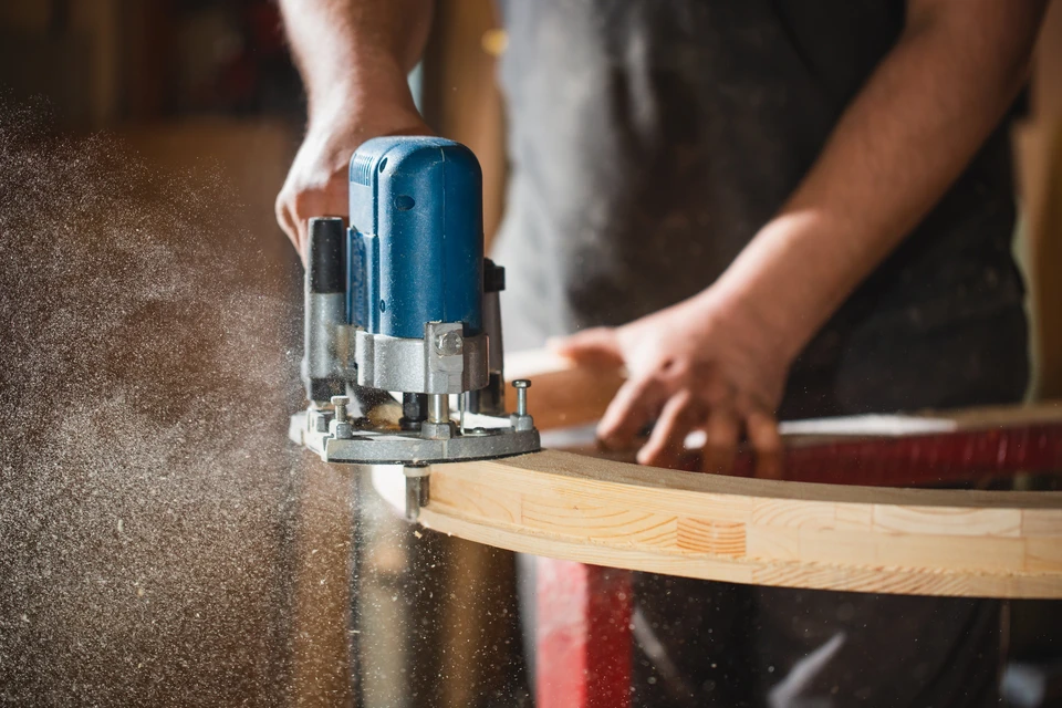 Atelier de menuiserie intérieur, artisan utilisant une défonceuse électrique bleue sur planche de bois massif