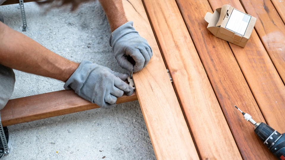 Un ouvrier qualifié en tenue professionnelle installant une terrasse bois composite sur un chantier résidentiel en extérieur, avec des éléments de fabrication (structure préassemblée, outillage professionnel) visibles à proximité, illustrant la transition entre fabrication et pose sur le terrain. Lumière naturelle, contexte résidentiel réaliste.