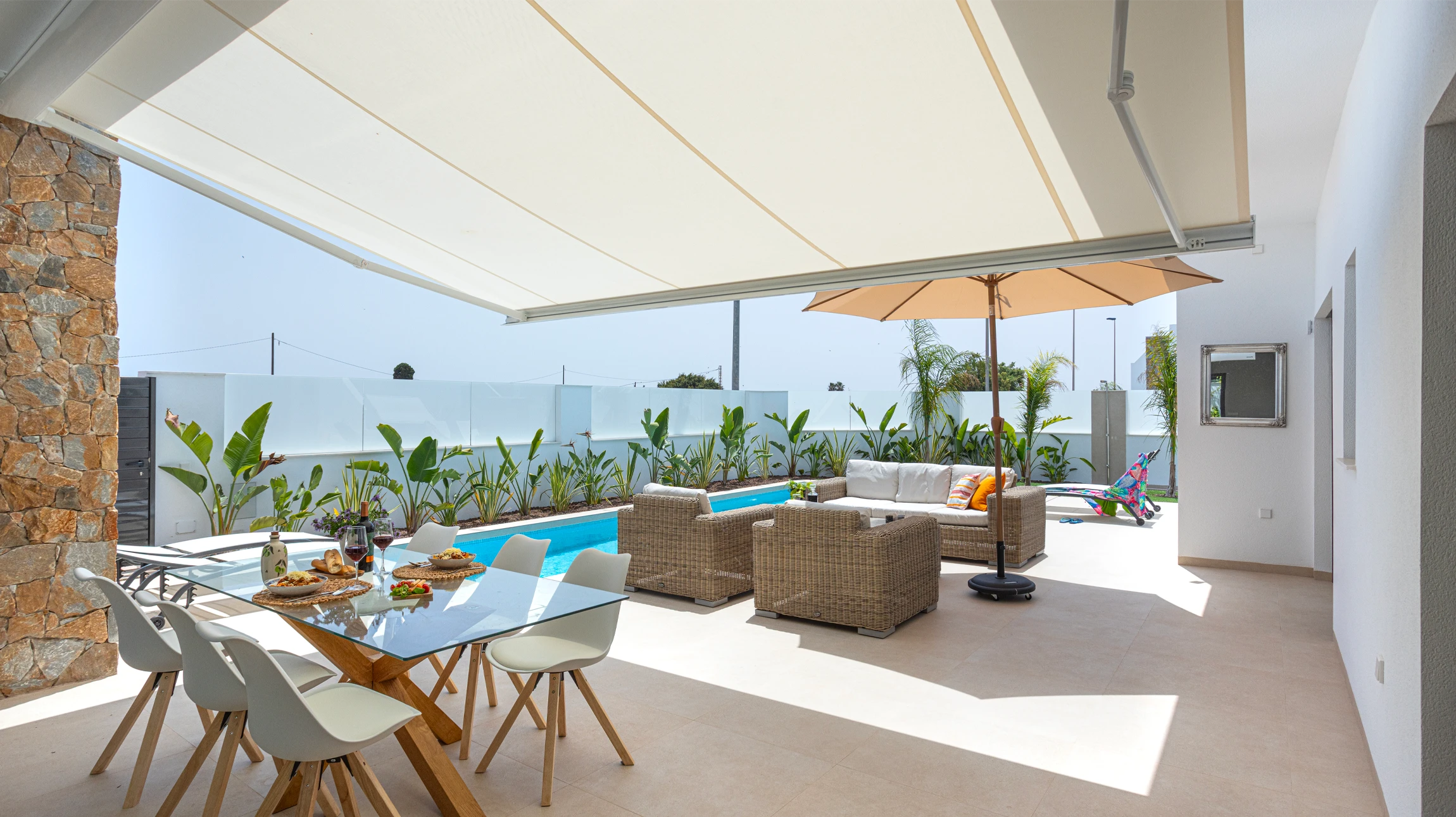 Photo réaliste d'une terrasse extérieure bien aménagée avec un store motorisé dépliée procurant l'ombre, créant un espace confortable. Vue de jour, montrant une personne détente depuis son canapé/fauteuil sous l'ombre du store, dans un jardin ou espace extérieur résidentiel agréable. L'accent doit être sur le confort et la détente permis par le système d'ombrage motorisé.