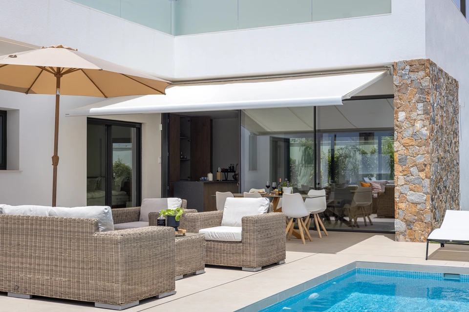 Intérieur lumineux d'une maison moderne avec une large baie vitrée ou fenêtre. Un store motorisé est partiellement fermé, filtrant la lumière naturelle de manière progressive. La photo capte la lumière tamisée créant une atmosphère confortable et contrôlée. La scène montre l'effet de protection solaire du store en action, avec des rayons de soleil filtrés créant des jeux de lumière doux sur le sol et les murs intérieurs. L'ambiance générale doit transmettre le confort, la sérénité et le contrôle intelligent de l'ensoleillement.