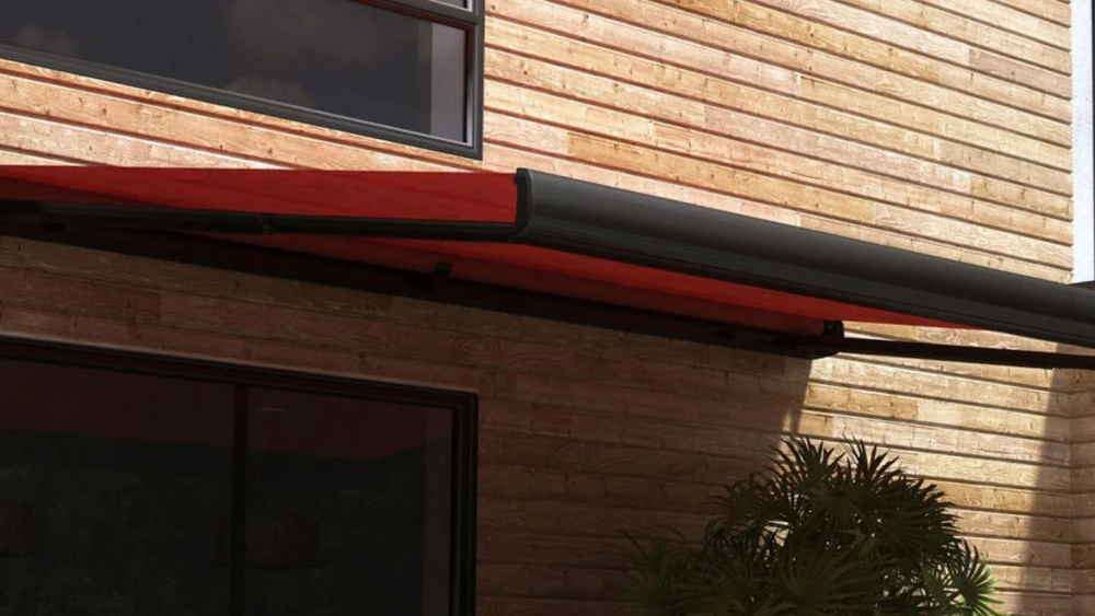 Photographie extérieure d'une pergola bioclimatique moderne en bois, montrant sa structure architecturale distinctive, avec lames orientables en position partiellement fermée, installée sur une terrasse résidentielle de style contemporain. La pergola crée une zone d'ombre élégante, bien intégrée à l'espace extérieur. Vue en contre-plongée ou angle latéral pour valoriser la structure et le design.