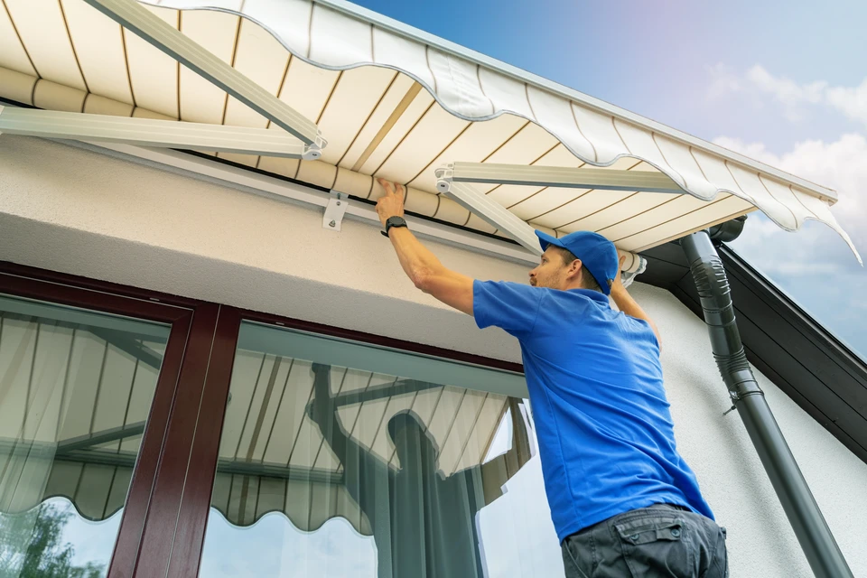 Artisan professionnel en tenue de travail, équipé de matériel de sécurité, en train d'installer un store motorisé extérieur vertical ou rétractable sur la façade d'une maison résidentielle. Le store, en tissu clair ou anthracite, est partiellement déroulé. L'installation se fait sur le mur extérieur de la maison, avec vue dégagée sur le paysage environnant. La scène capture un moment de travail réel sur chantier, montrant le professionnalisme et la précision de l'installation d'un produit d'aménagement extérieur.