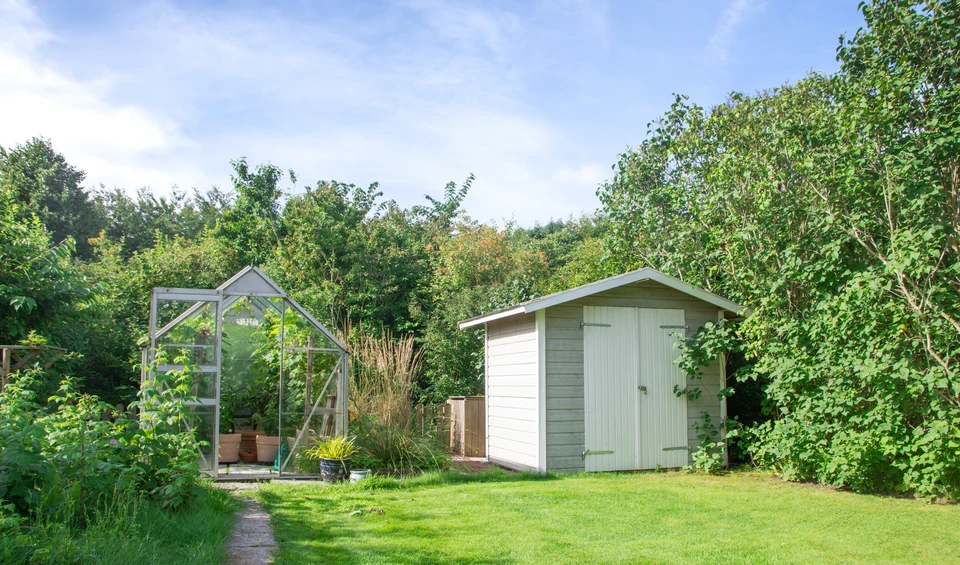 Photographie d'un abri de jardin en bois clair ou en bois composite, de style moderne et épuré, construit dans un jardin paysagé. L'abri est légèrement ouvert ou partiellement visible, montrant son intérieur organisé avec des étagères en bois contenant des outils de jardinage, des pots de fleurs, des équipements de jardinage rangés de manière fonctionnelle. Le bâtiment s'intègre harmonieusement dans le jardin, avec du mobilier d'extérieur à proximité (banc ou salon de jardin discret). La photo est prise en lumière naturelle mettant en évidence la qualité de la construction et la praticité de l'espace de rangement.