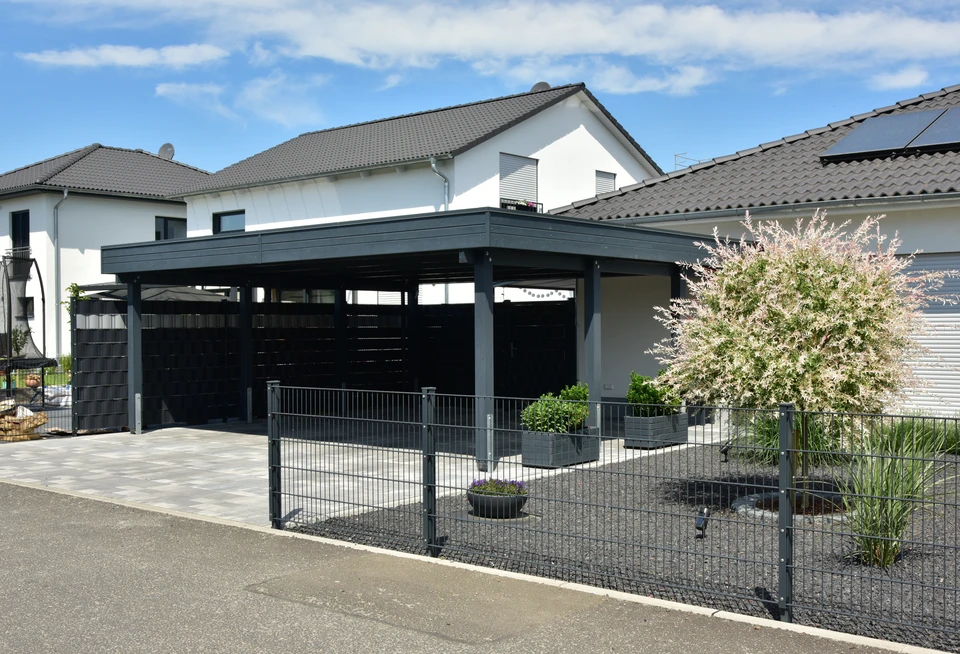 Photographie d'un carport en bois ou aluminium de construction moderne, implanté dans un espace extérieur résidentiel typique. Le carport abrite un véhicule de couleur claire visible dessous, démontrant sa fonctionnalité. La structure est bien intégrée à l'environnement de la maison, avec une pelouse et éventuellement un jardin adjacent. La photo est prise de jour, avec une bonne luminosité naturelle, montrant la finition et la qualité de la construction. L'accent est mis sur l'élégance et la fonctionnalité de la structure.