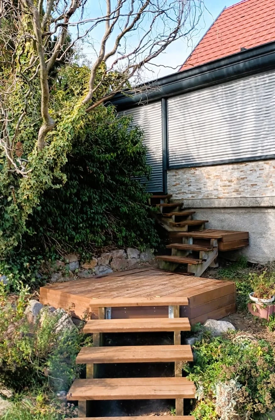 Terrasse en bois surélevée fixée en façade d'un immeuble résidentiel gris, accessible par un escalier métallique latéral